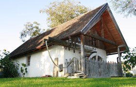 Museum außen, © Fam. Buchinger Traditionelles Gebäude mit Holzdach und Zaun, umgeben von grüner Wiese und Bäumen.