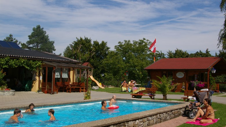 Cool by the pool, © Reinhard Wallentin People relax by a small pool in a garden with wooden houses and a slide.