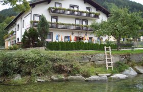 Gasthof Lichtensteg, © Archiv Betrieb Ein traditionelles Gasthaus mit Balkon und Blumenkästen, umgeben von Bäumen und einem Fluss im Vordergrund.