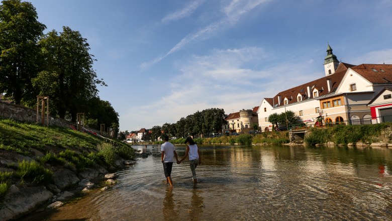 Erlaufhafen, © schwarz-koenig.at Ein Paar geht Hand in Hand durch einen flachen Fluss in einer Stadt mit historischen Gebäuden und Bäumen.