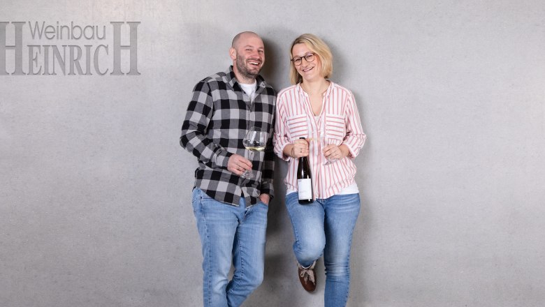 Holder, © Weinbau Heinrich Two people stand smiling in front of a gray wall with wine glasses in their hands. On the left is a man, on the right a woman. At the top left it says 'Weinbau Heinrich'.
