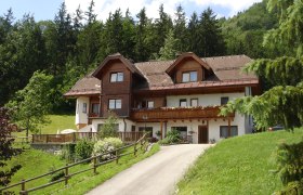 Haus Esletzbichler, © zVg Ein traditionelles Haus mit Holzverkleidung in einer grünen, bewaldeten Umgebung.