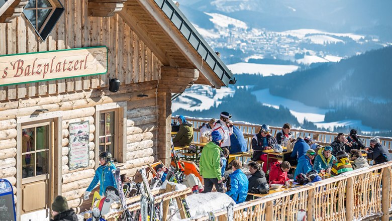 Sonnenterrasse s'Balzplatzerl, © Martin Krcal Menschen auf einer sonnigen Terrasse vor einer Berghütte mit Blick auf verschneite Berge.