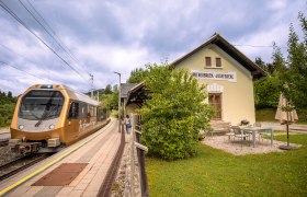 Urlaub am BAHNhof, © Niederösterreich Werbung / Maximilian Pawlikowsky Ein goldener Zug hält am Bahnhof Wienerbruck-Josefsberg, umgeben von grüner Landschaft und einem kleinen Sitzbereich im Freien.