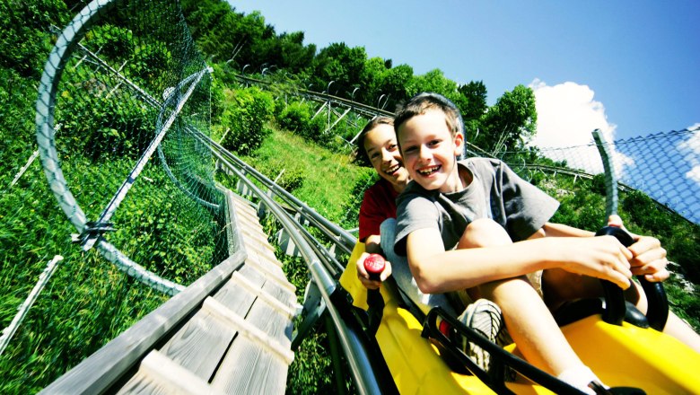 Allwetter-Rodelbahn Eibl-Jet in Türnitz, © weinfranz.at Zwei Kinder fahren lachend auf einer Sommerrodelbahn durch grüne Landschaft.