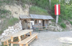 Wohnhöhle, © ZVG Gemeinde Eine rustikale Holzhütte mit Tisch und Bänken davor, eingebaut in einen Hügel. Eine rot-weiße Fahne hängt an einem Baum.