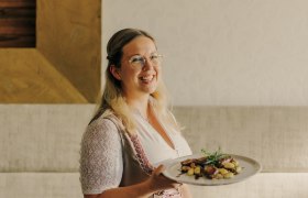 Wirtin Sabrina Stadler, © Niederösterreich Werbung/Julius Hirtzberger Frau in traditioneller Kleidung serviert ein Gericht auf einem Teller.