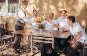 Starkes Wirtshaus-Team, © Niederösterreich Werbung/Daniela Führer Ein Team von fünf Personen stößt draußen an einem Tisch an, mit einem schwarzen Hund im Vordergrund.