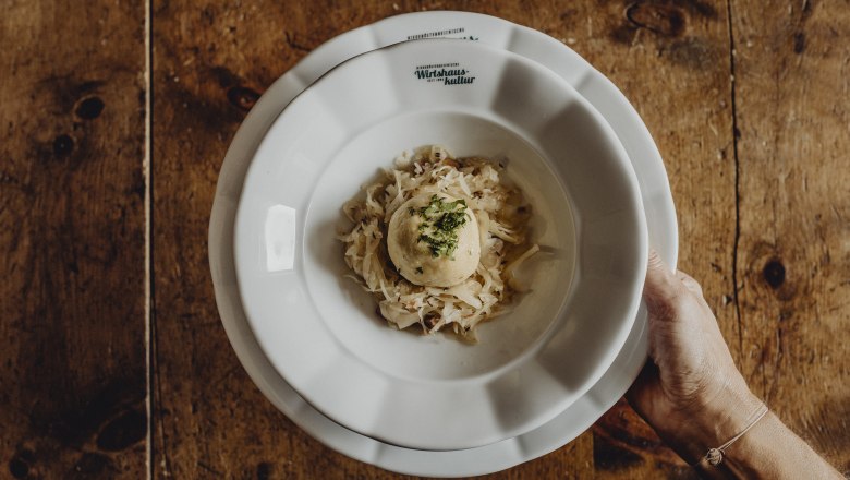 Grammelknödel, © Niederösterreich Werbung/Sophie Menegaldo Ein Teller mit einem Grammelknödel auf Sauerkraut, gehalten von einer Hand.