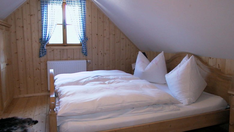 Zimmer, © Ferienhaus Hintereck Gemütliches Schlafzimmer mit Holzwänden, einem Doppelbett und einem Fenster mit blau karierten Vorhängen.