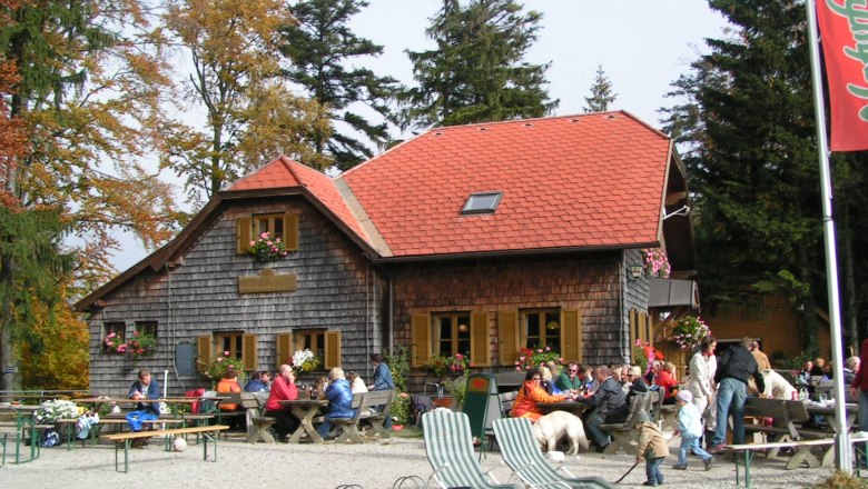 Ochsenburger Hütte, © Naturfreunde St. Georgen Eine Berghütte mit rotem Dach und Holzwänden, umgeben von Bäumen. Menschen sitzen draußen an Tischen, einige mit Hunden.