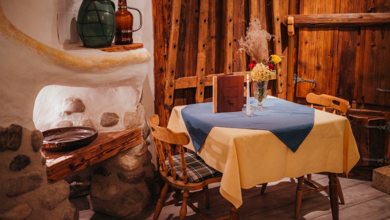 Seit 1977 ist der Bauernhof ein Gasthaus, © Niederösterreich Werbung/Daniela Führer Rustikales Gasthausinterieur mit Holzmöbeln, Tisch mit gelber Tischdecke, blauer Serviette, Blumen und Kerze.