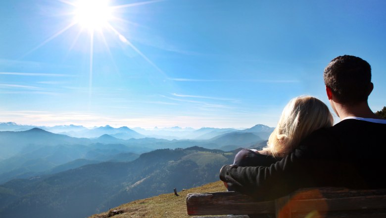 Ausblick von der Gemeindealpe, © weinfranz.at Ein Paar sitzt auf einer Bank und blickt auf eine Berglandschaft unter einem strahlend blauen Himmel mit Sonne.