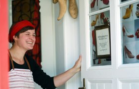 Doris Pfaffenlehner, © weinfranz.at Eine Frau mit rotem Stirnband und Schürze steht lächelnd an einer Tür mit Schuhbildern und einem Schild der Maßschuhmacherei Pfaffenlehner Doris.