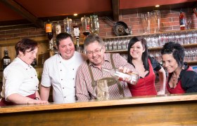 Schliefauhof, © weinfranz.at Eine Gruppe von Menschen hinter einer Bar, die gemeinsam lachen und eine Flasche in einen Krug gießen.