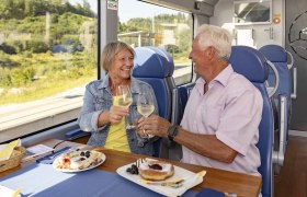 Mariazellerbahn, © NÖVOG/Schwarz-König Ein älteres Paar stößt mit Weingläsern in einem Zug an, mit Essen auf dem Tisch.