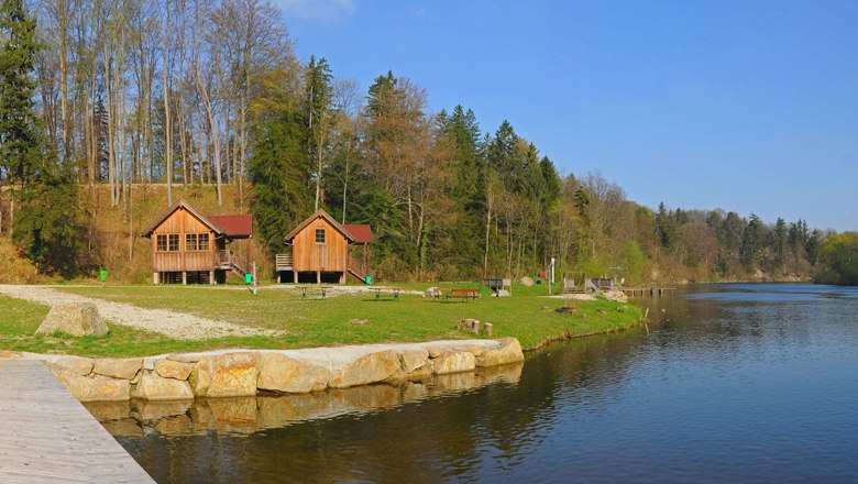 Natural swimming pool Kematen, © zVg Gemeinde Kematen Two wooden huts on the shore of a lake, surrounded by trees and green grass.