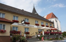 Ein traditioneller Gasthof mit Blumen geschm&uuml;ckter Fassade und Terrasse, daneben eine Kirche mit spitzem Turm.