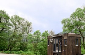 Ein kleiner, dunkler Wohnwagen mit Holzverkleidung steht auf einem Parkplatz vor gr&uuml;nen B&auml;umen und bew&ouml;lktem Himmel.