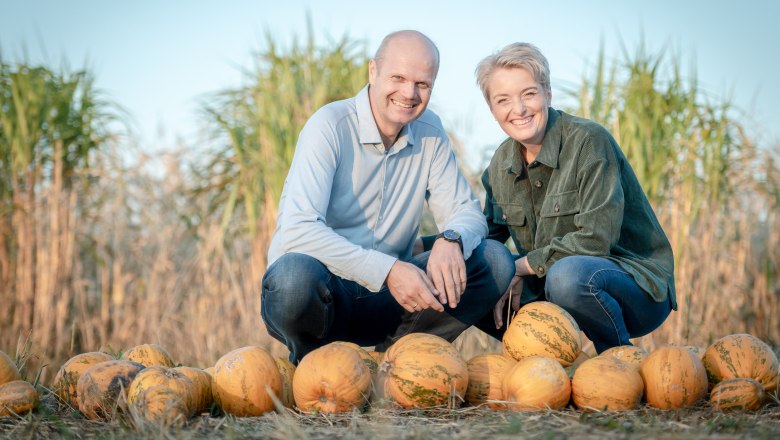 Two people kneel smiling behind a row of pumpkins in a field.