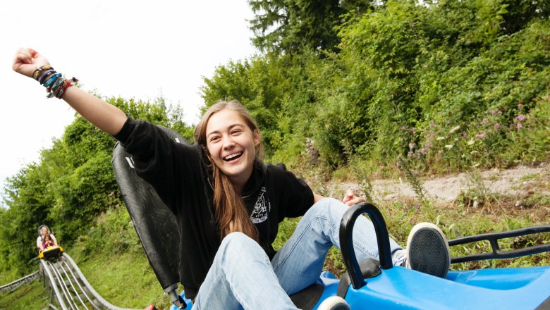 Eine lachende Person f&auml;hrt auf einer Sommerrodelbahn, umgeben von gr&uuml;ner Natur.