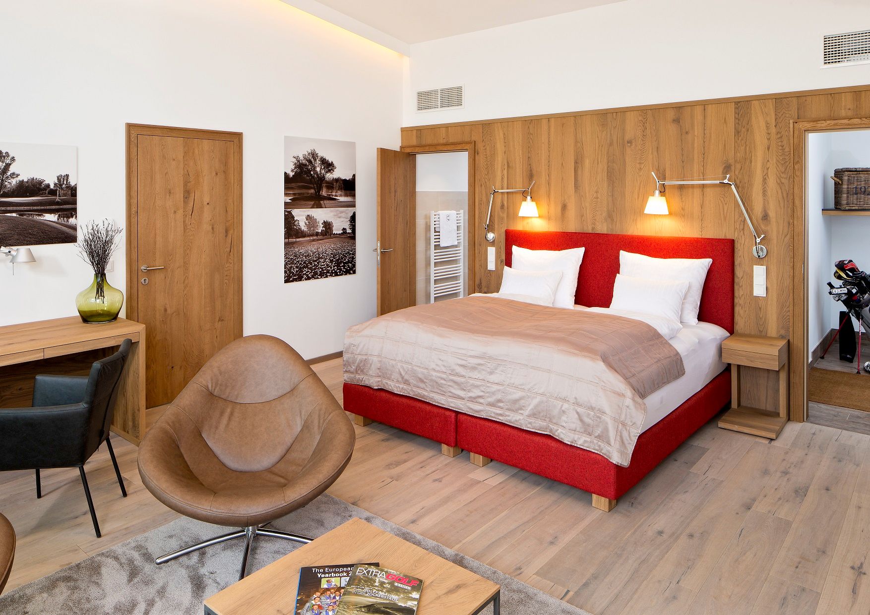 Modernes Hotelzimmer mit rotem Bett, Holzmöbeln und Golfmagazinen auf dem Tisch.