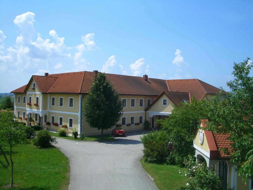 Großer Bauernhof mit gelben Wänden und roten Dächern, umgeben von Bäumen und einer Straße.