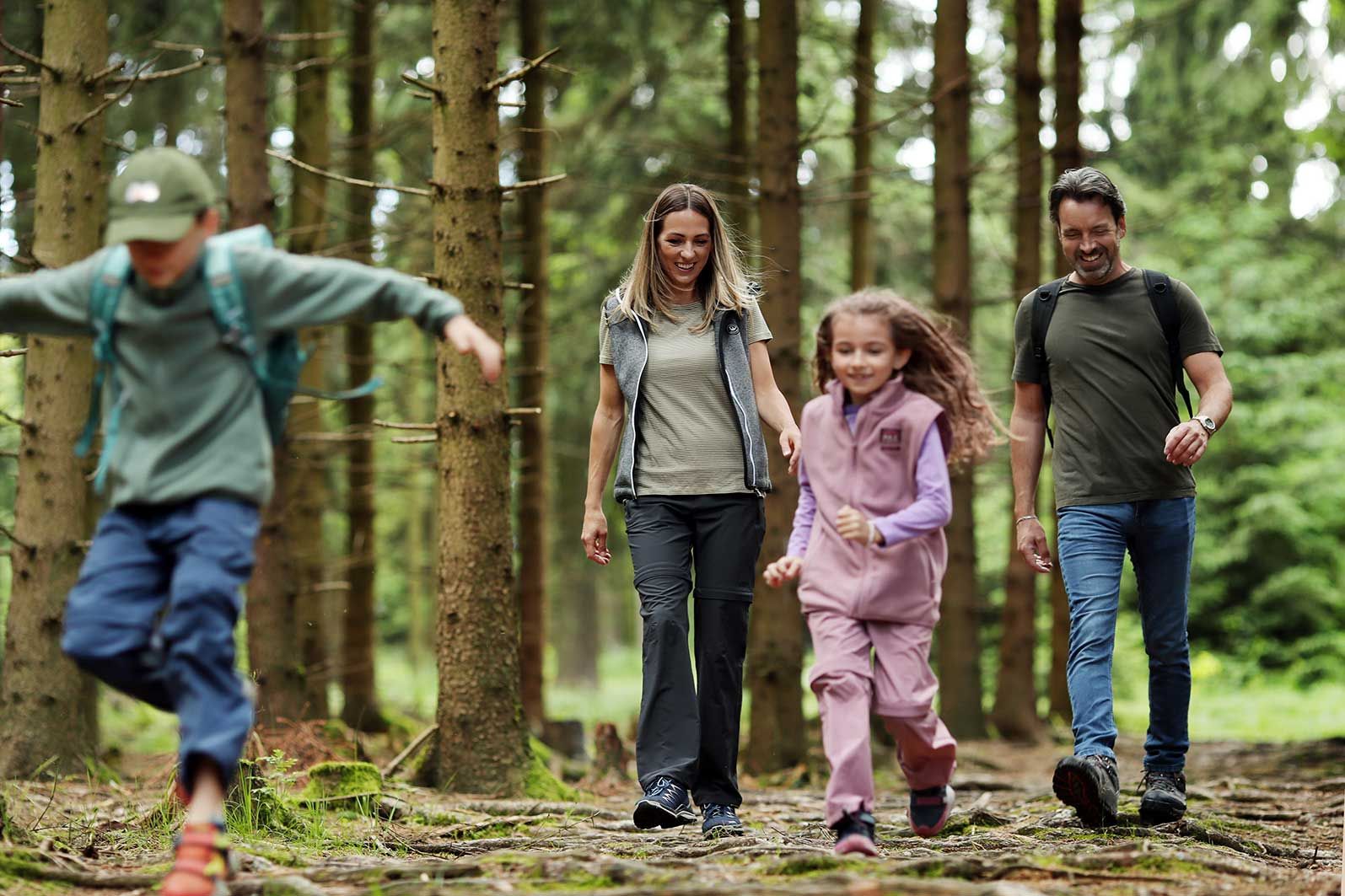 Eine Familie mit einem Jungen und einem Mädchen wandert in Wanderkleidung durch einen Fichtenwald. Der Junge im Vordergrund springt. Im Hintergrund ist eine Lichtung.