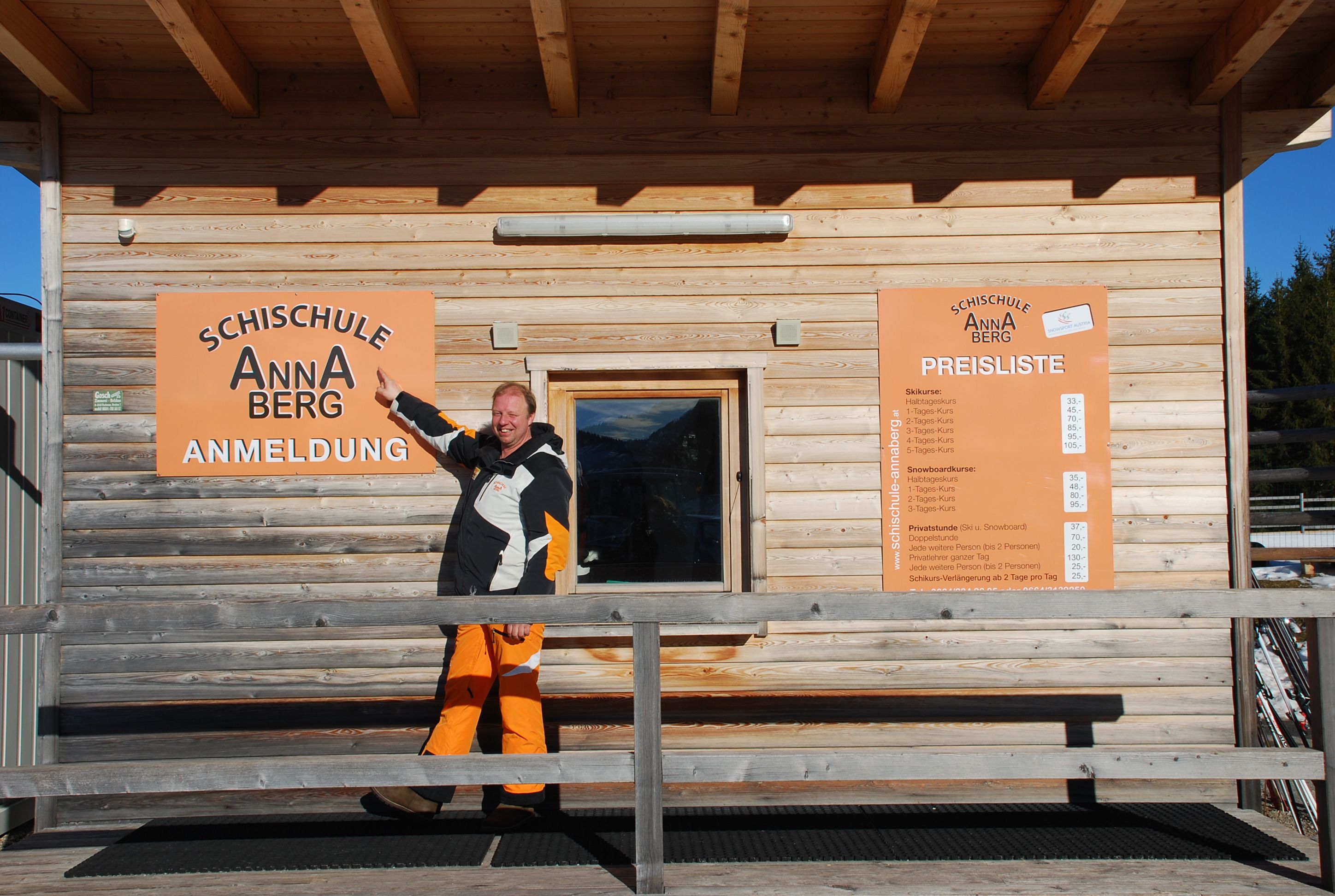 Ein Mann zeigt auf ein Schild der Schischule Annaberg vor einem Holzgebäude.