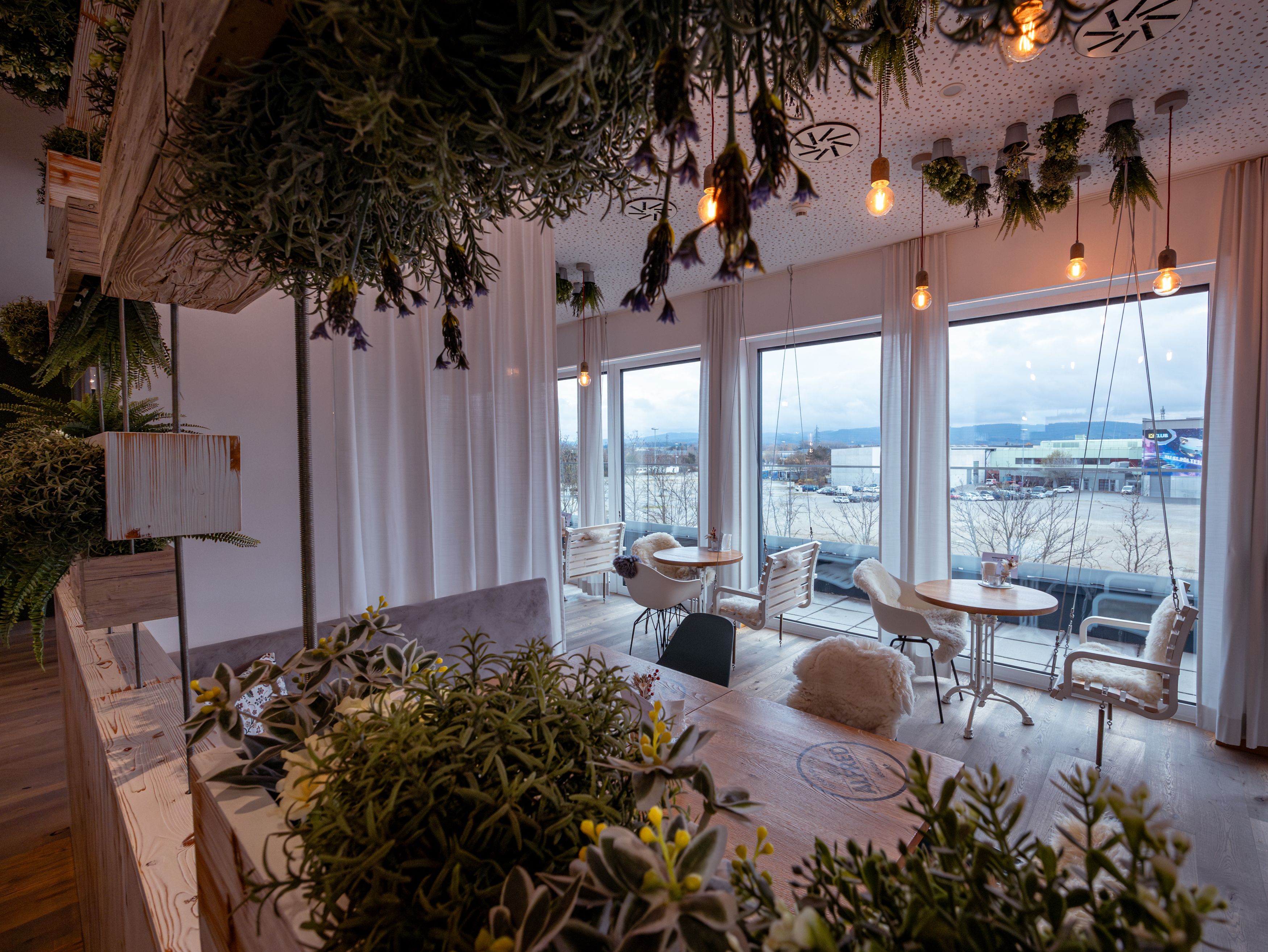 Innenraum eines Cafés mit Pflanzen und großen Fenstern.