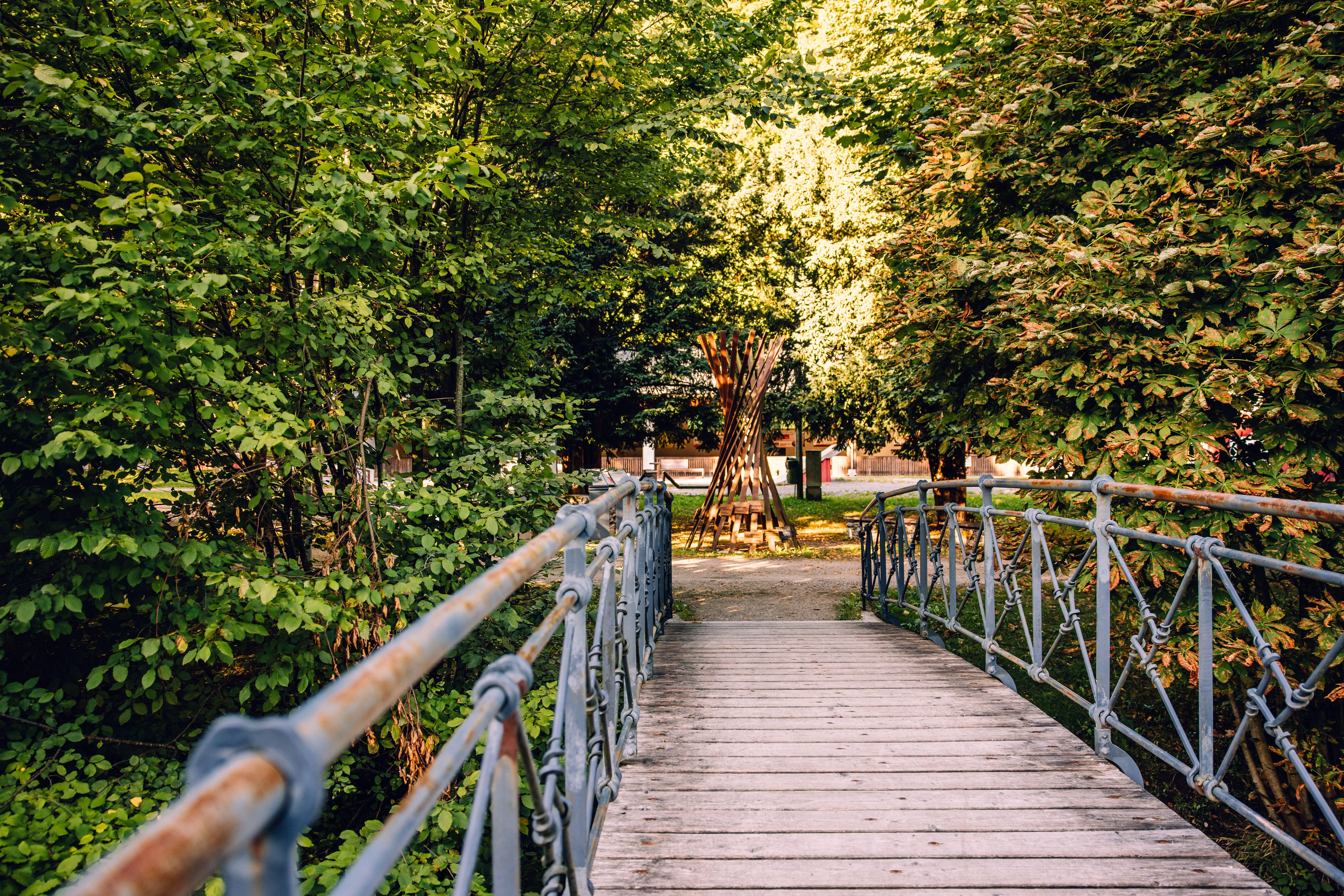 Holzbrücke mit kunstvoll geschmiedetem Metallgeländer entlang der Schmiedemeile in Ybbsitz, umgeben von grünen Bäumen und mit Blick auf eine Holzskulptur.