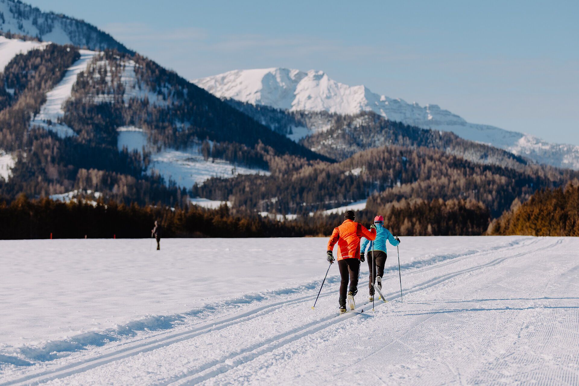 Die glitzernde Schneedecke erstreckt sich über die sanften Hügel, während zwei Langläufer in lebhaften Farben durch die winterliche Landschaft gleiten. Die klare, kalte Luft und die majestätischen Berge im Hintergrund schaffen eine perfekte Kulisse für unvergessliche Erlebnisse im Schnee.