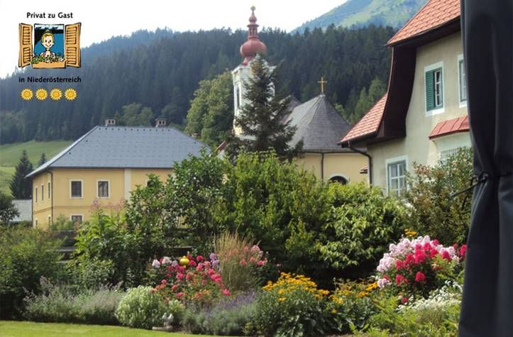 Ein malerischer Garten mit bunten Blumen vor traditionellen Häusern und einer Kirche in einer bergigen Landschaft.