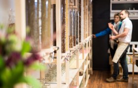 Innenansicht eines Unverpackt-Ladens mit Spendern für lose Lebensmittel und zwei Personen im Hintergrund.