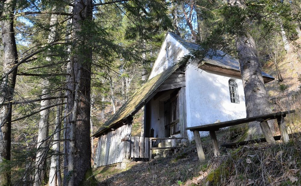 A small white chapel in the forest, surrounded by trees and a wooden table in the foreground.
