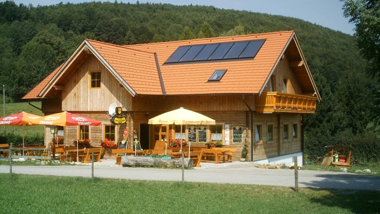 Ein traditionelles Gasthaus mit Holzw&auml;nden und roten Dachziegeln, umgeben von gr&uuml;ner Landschaft und Wald.
