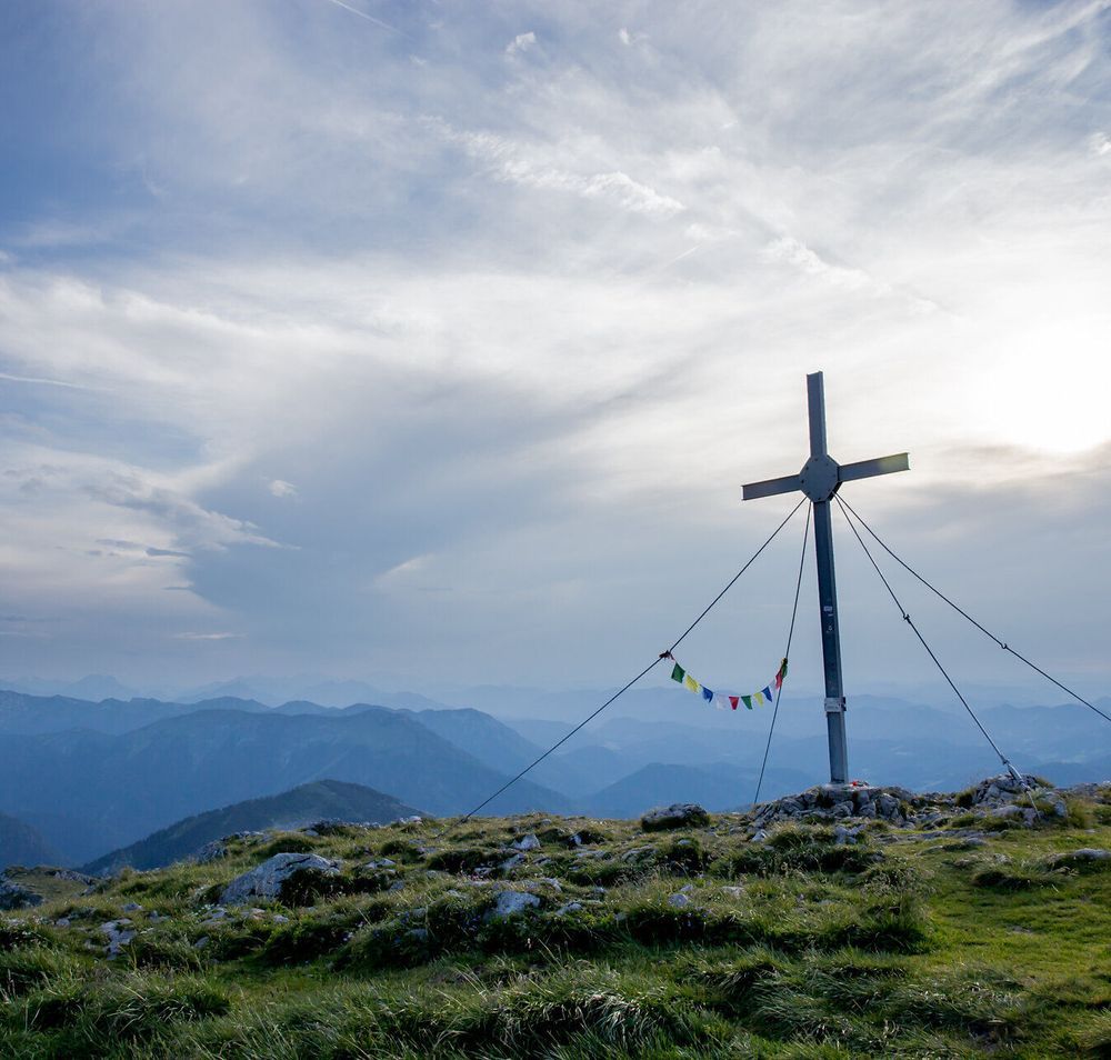 A majestic summit cross towers over the rolling hills, surrounded by a breathtaking mountain landscape. The clouds gently drift across the sky and lend the scenery a peaceful atmosphere, while the fresh mountain air invites you to go hiking.