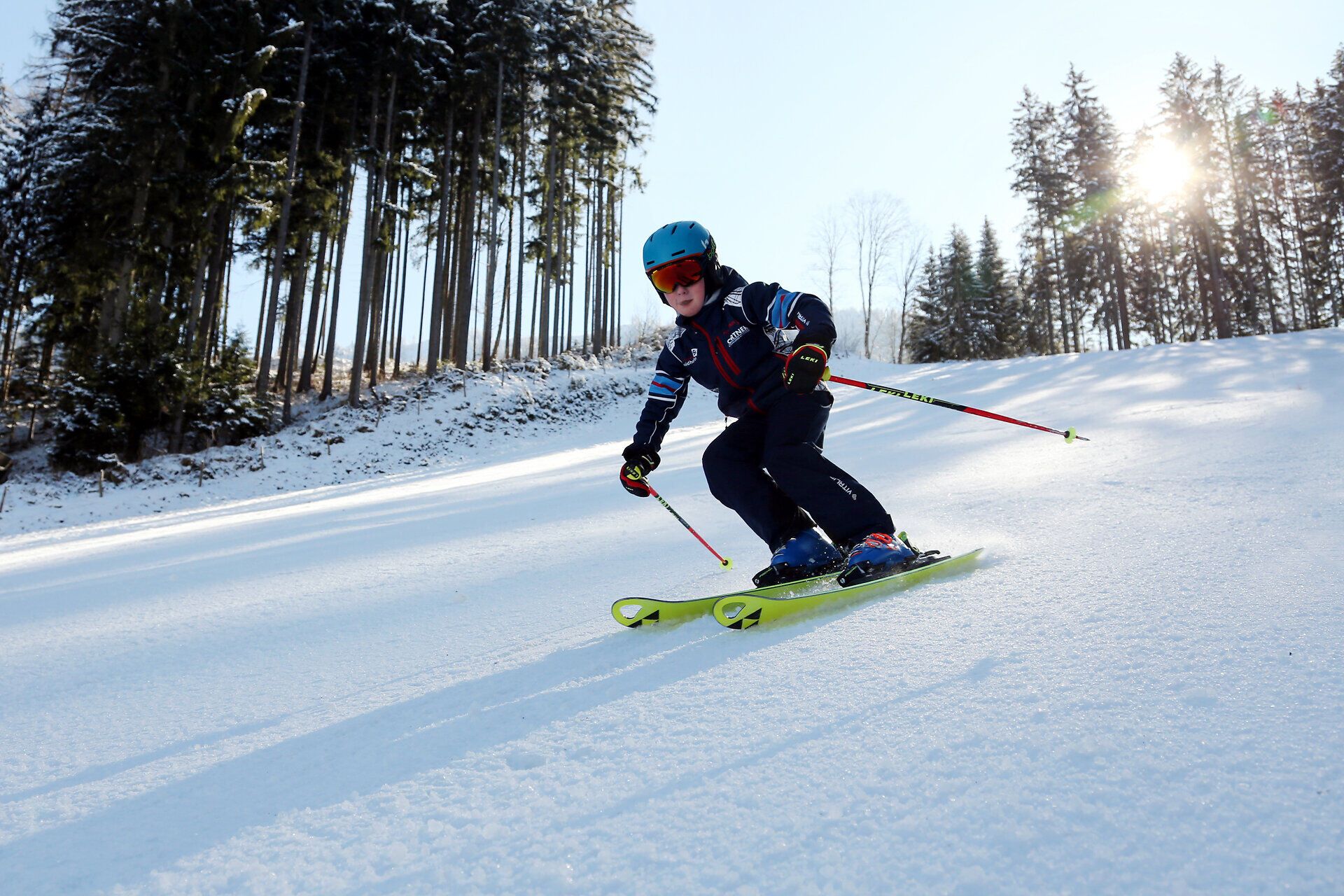Ein strahlend blauer Himmel umrahmt die schneebedeckte Piste, während ein fröhliches Kind mit Begeisterung die Abfahrt hinuntergleitet. Die frische Bergluft und die glitzernde Schneedecke schaffen eine unvergessliche Winteratmosphäre, die Familien zu gemeinsamen Abenteuern einlädt.
