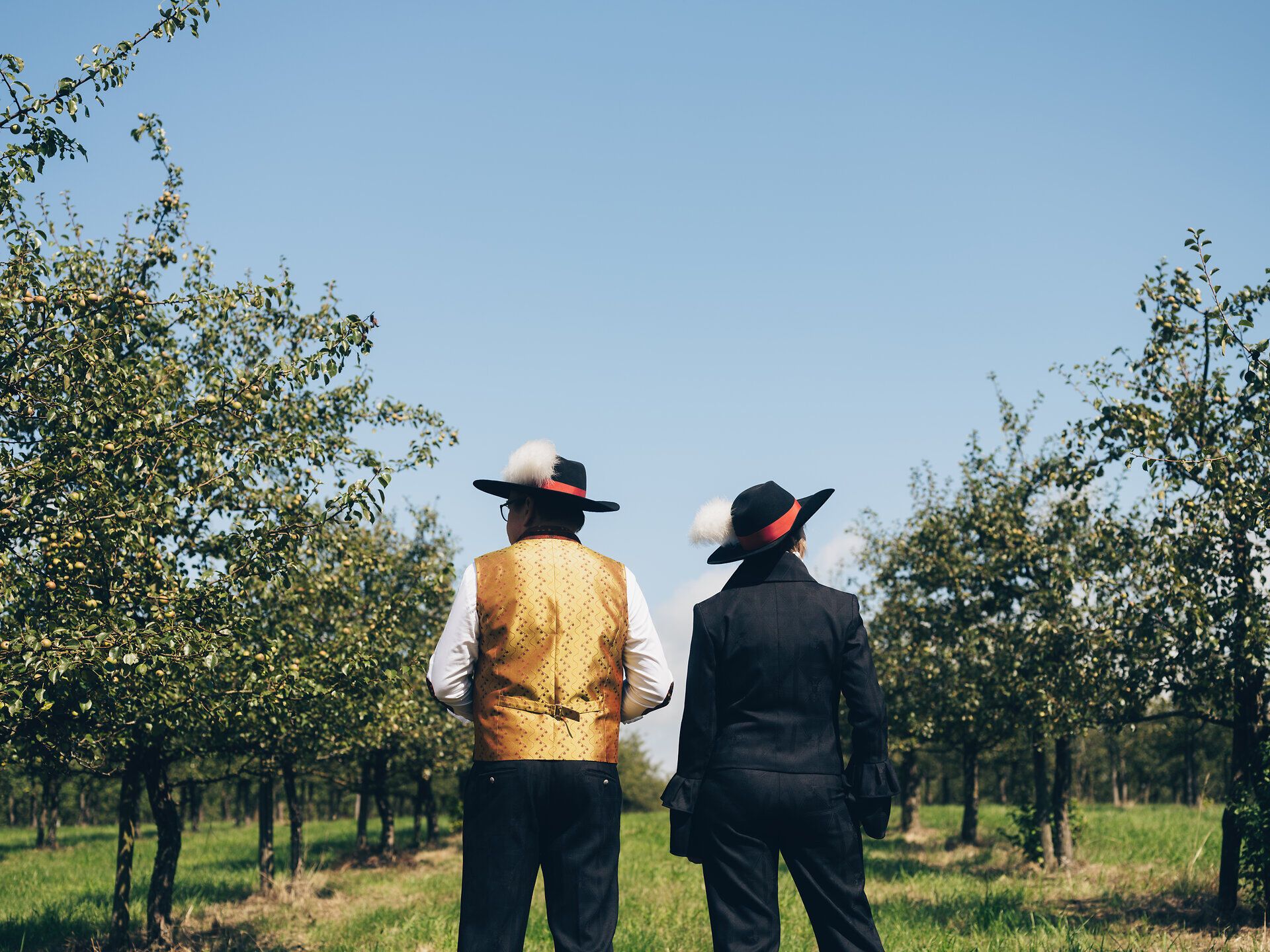 Inmitten der sanften Hügel des Mostviertels stehen zwei Personen in traditioneller Tracht und genießen die malerische Aussicht auf die Birnenbäume, die in voller Blüte stehen. Die klare Luft und das sanfte Licht des Sommertages schaffen eine einladende Atmosphäre, die zum Verweilen einlädt.