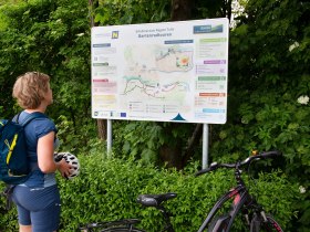 Žena s cyklistickou přilbou a batohem na z&aacute;dech si prohl&iacute;ž&iacute; informačn&iacute; tabuli o zahradn&iacute;ch cyklistick&yacute;ch v&yacute;letech v regionech Tulln obklopen&yacute;ch zelen&iacute;.