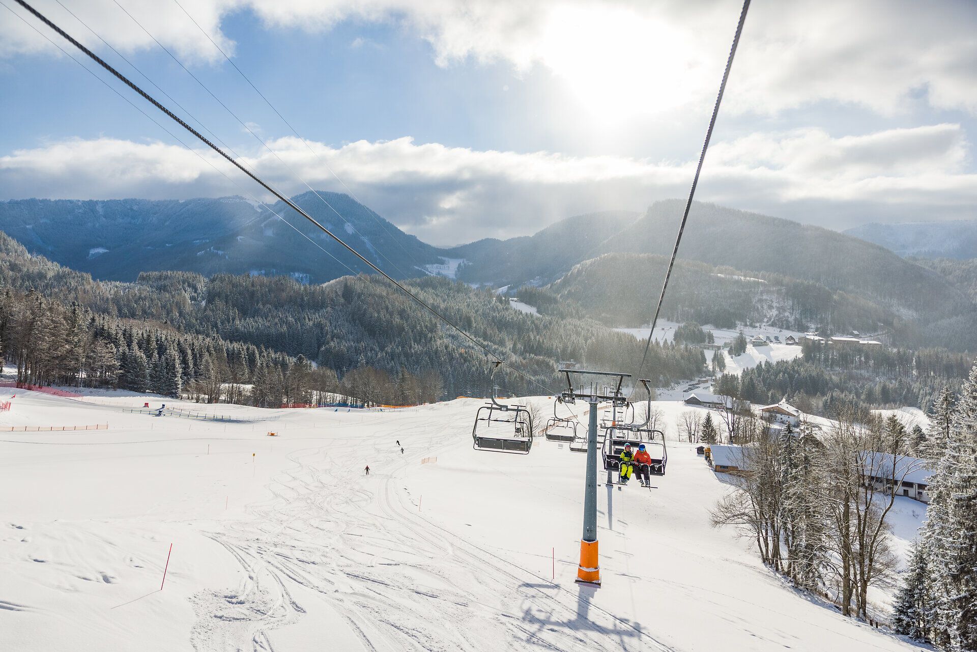 Die glitzernde Schneedecke erstreckt sich über die sanften Hügel, während der Sessellift gemächlich die Skifahrer zu neuen Abenteuern hinaufträgt. Die klare, kalte Luft und die majestätischen Berge schaffen eine perfekte Kulisse für unvergessliche Wintertage.