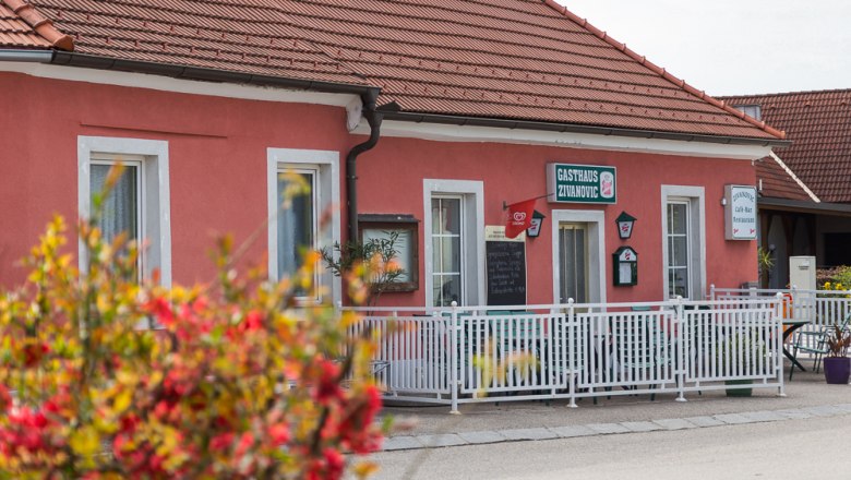 A pink building with the inscription 'Gasthaus Zivanovic', surrounded by a white fence and flowers in the foreground.