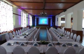 Festlich gedeckter Saal mit langen Tafeln und Projektionsleinwand.