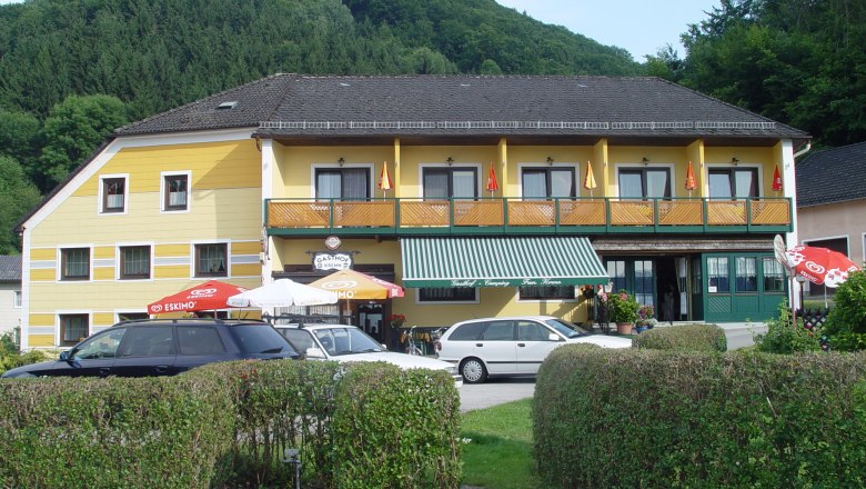 Gelbes Gasthaus mit Balkon und Sonnenschirmen vor einem bewaldeten Hügel.