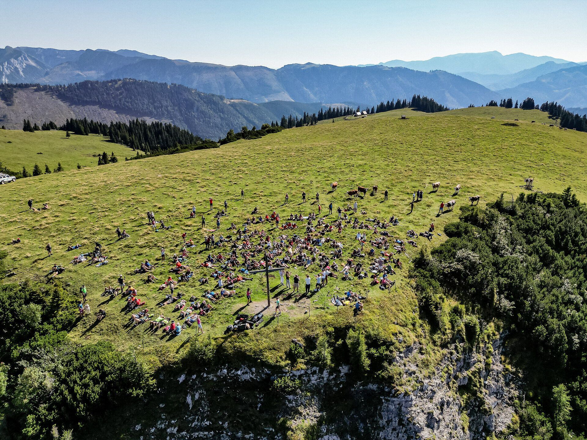 Inmitten der sanften Hügel und der beeindruckenden Berglandschaft versammeln sich zahlreiche Menschen, um die Klänge der Natur und Musik zu genießen. Die grüne Wiese bietet einen perfekten Platz für Entspannung und Geselligkeit, während die majestätischen Berge im Hintergrund eine atemberaubende Kulisse schaffen.