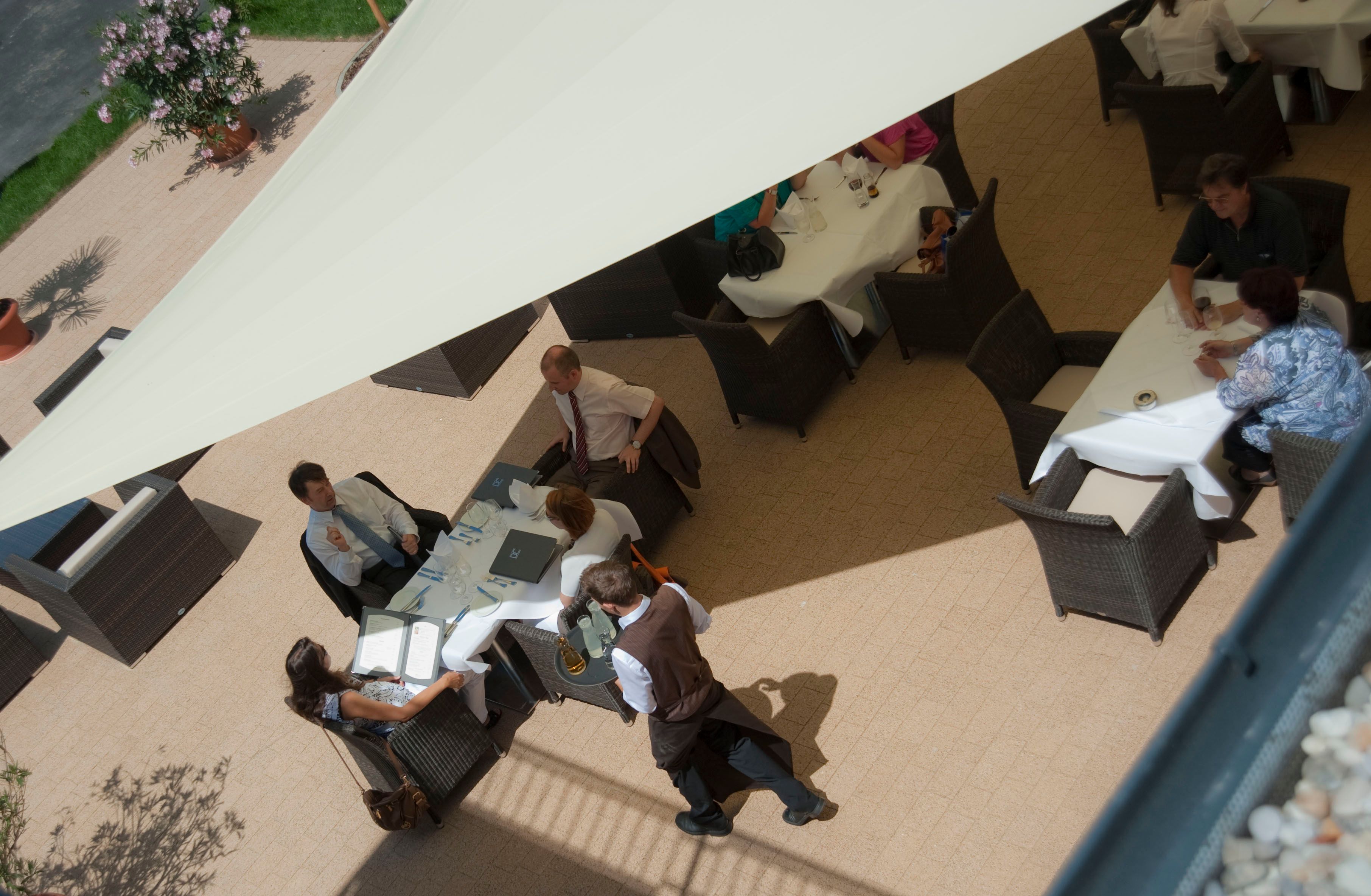 Luftaufnahme einer Hotelterrasse mit Gästen an Tischen unter einem Sonnensegel.
