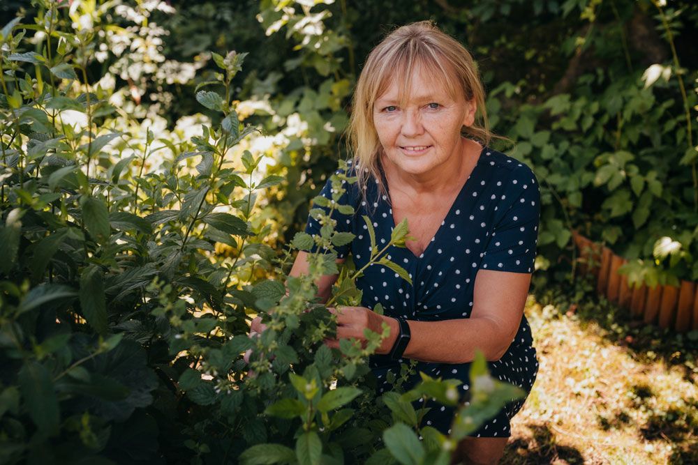Eine Frau in einem gepunkteten Kleid kniet in einem Garten und betrachtet Pflanzen.