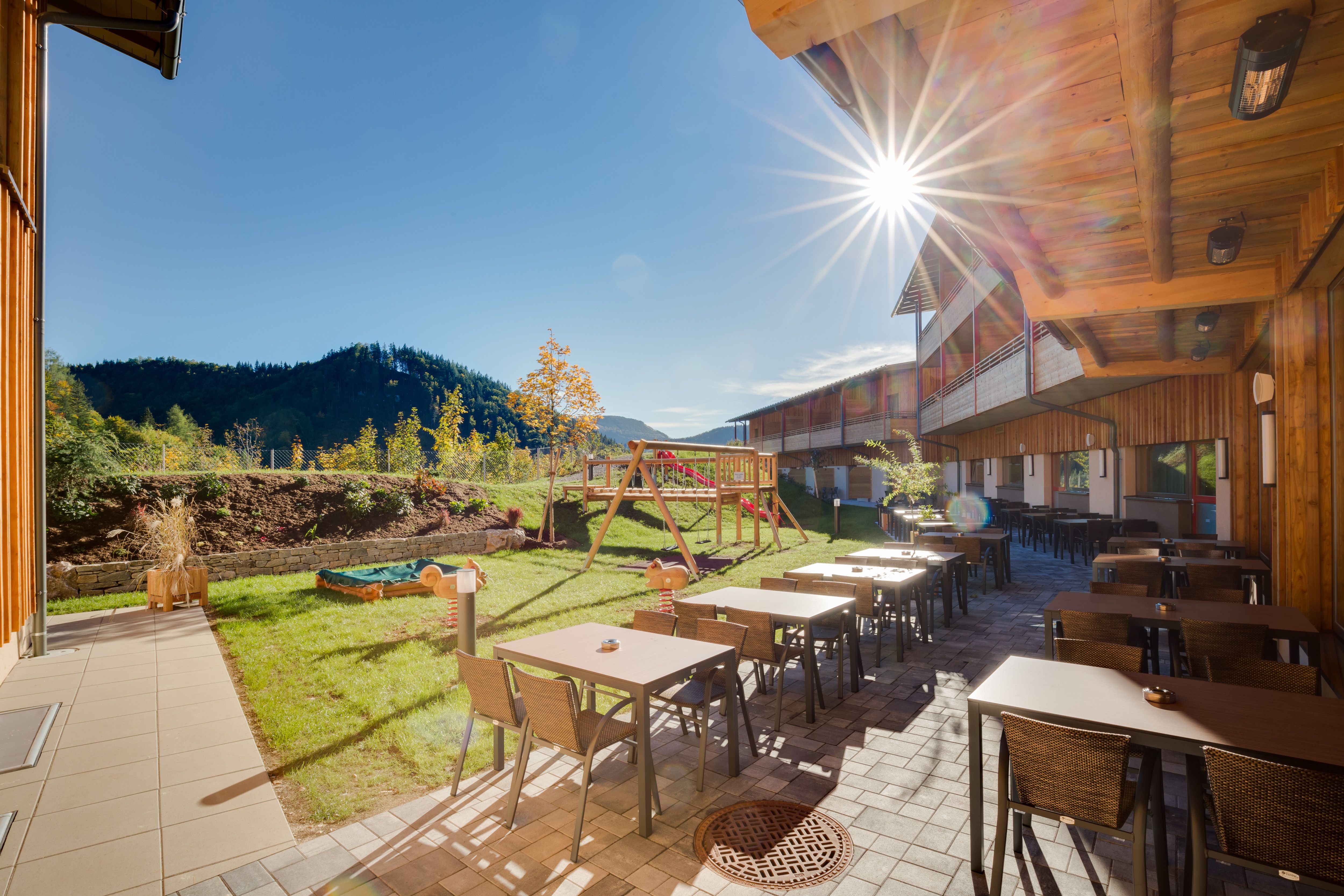 Terrasse des JUFA Hotels Annaberg mit Tischen, Stühlen und Spielplatz im Hintergrund bei Sonnenschein.
