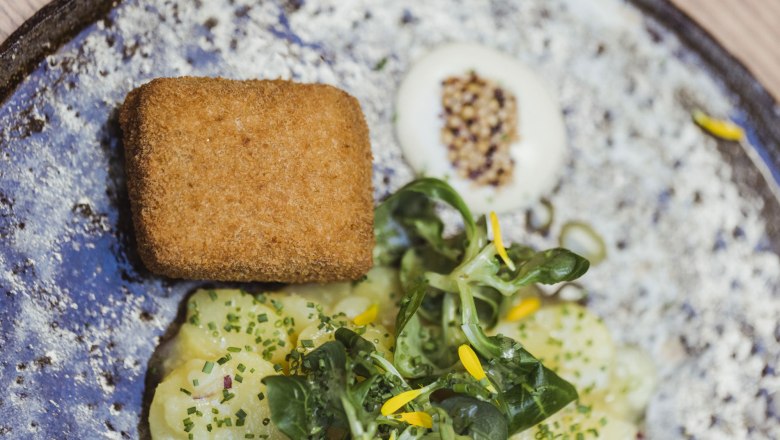 Ein Teller mit paniertem Kalbskopf, Kartoffelsalat und Feldsalat, garniert mit gelben Blüten.
