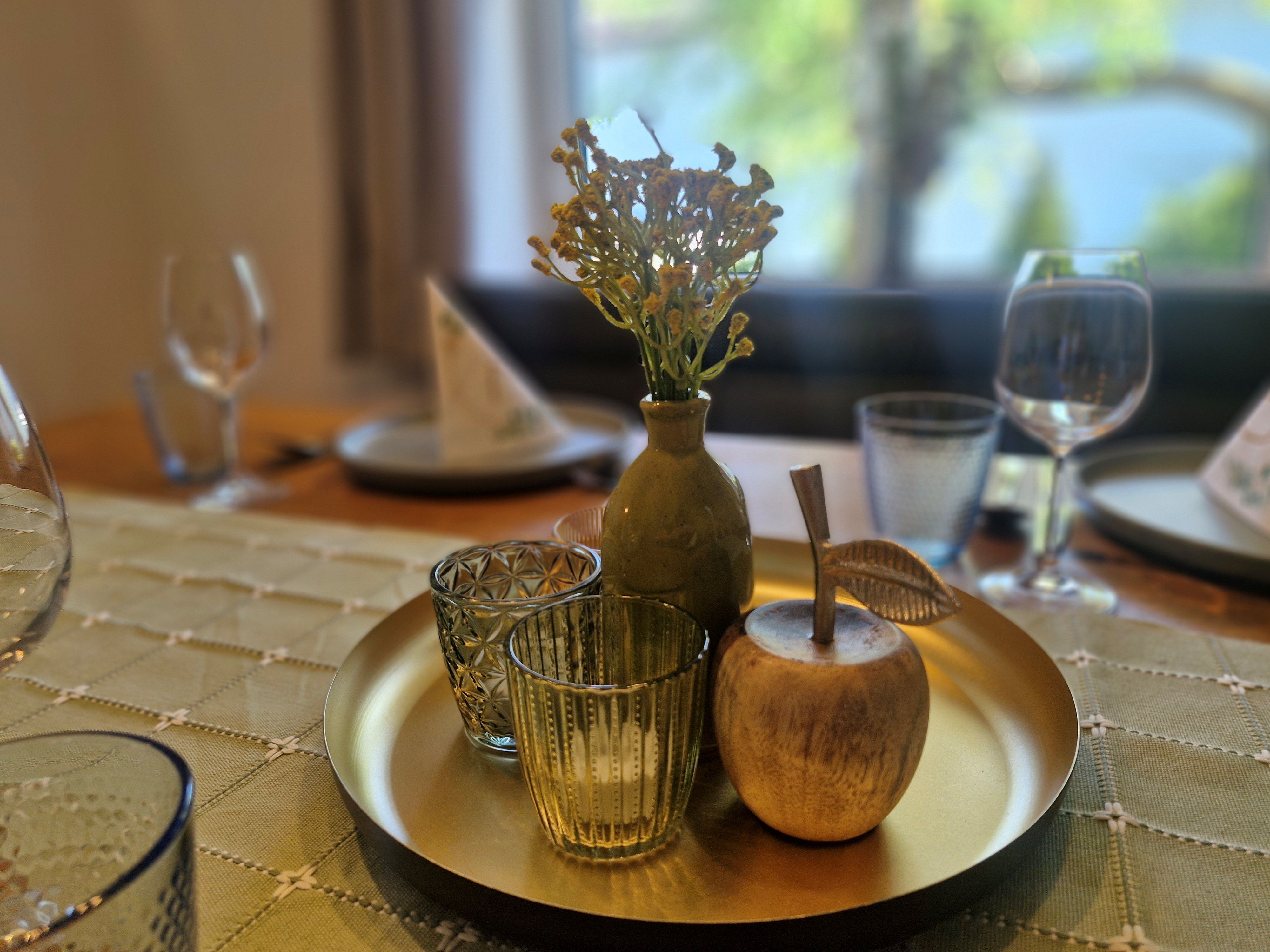 Ein dekorativer Tisch mit einem goldenen Tablett, auf dem eine Vase mit Blumen, zwei Gläser und ein hölzerner Apfel stehen.